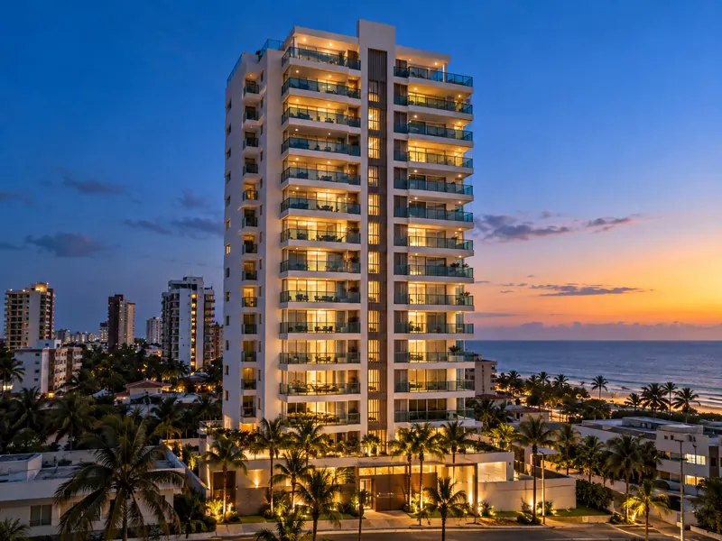 Torre residencial branca com varandas iluminadas ao entardecer, perto do mar